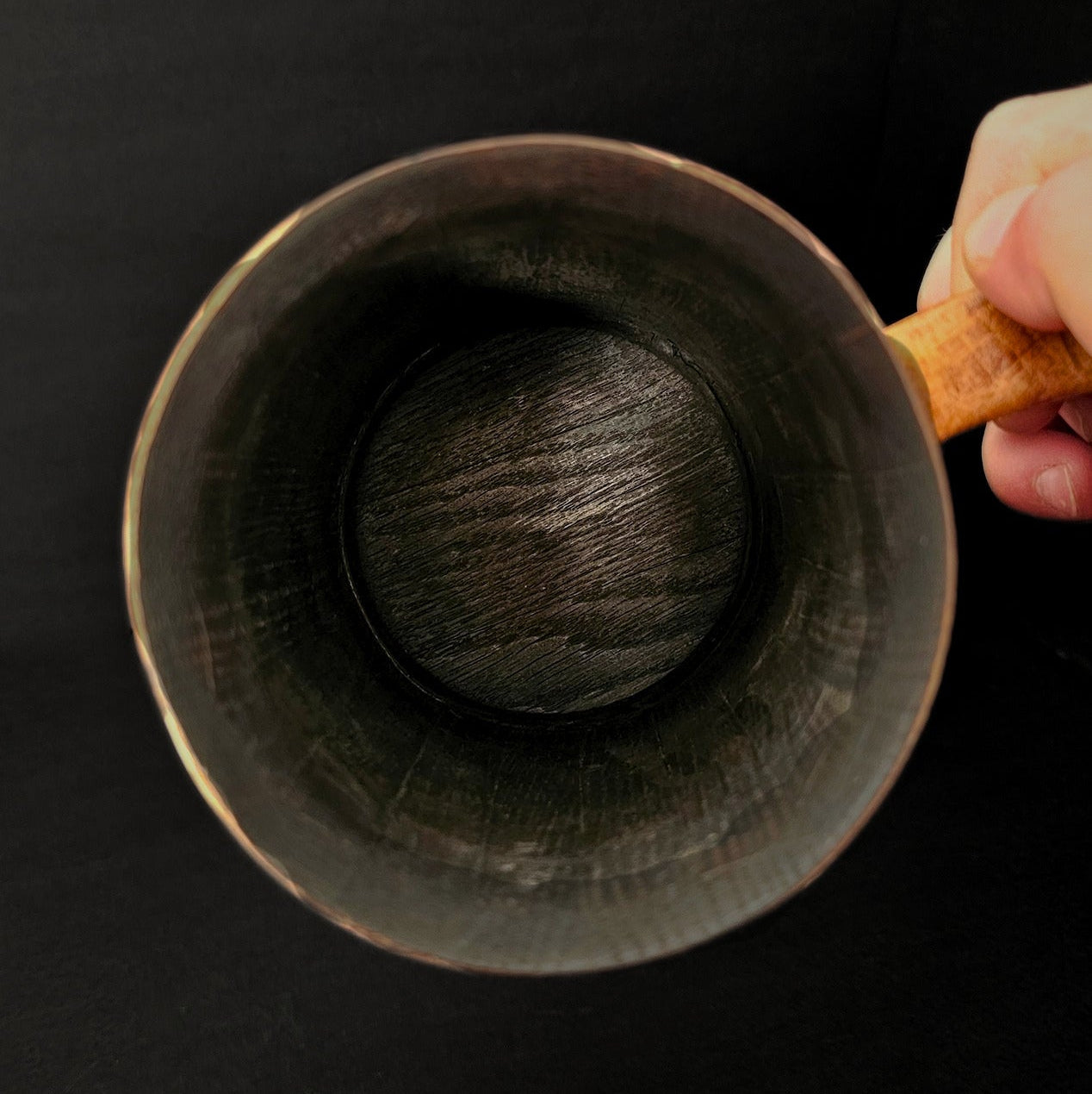 Hand holding oak tankard to show inside of tankard. Charring visible. Black background.